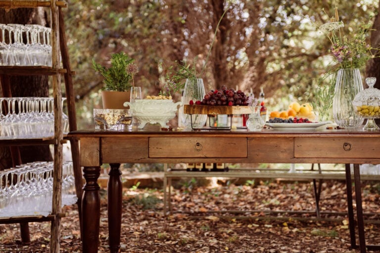 tavolo vintage con cassetti in primo piano all'aperto apparecchiato con uva, frutti di bosco, olive decorato con fiori, sulla sinistra un mobile porta calici con calici