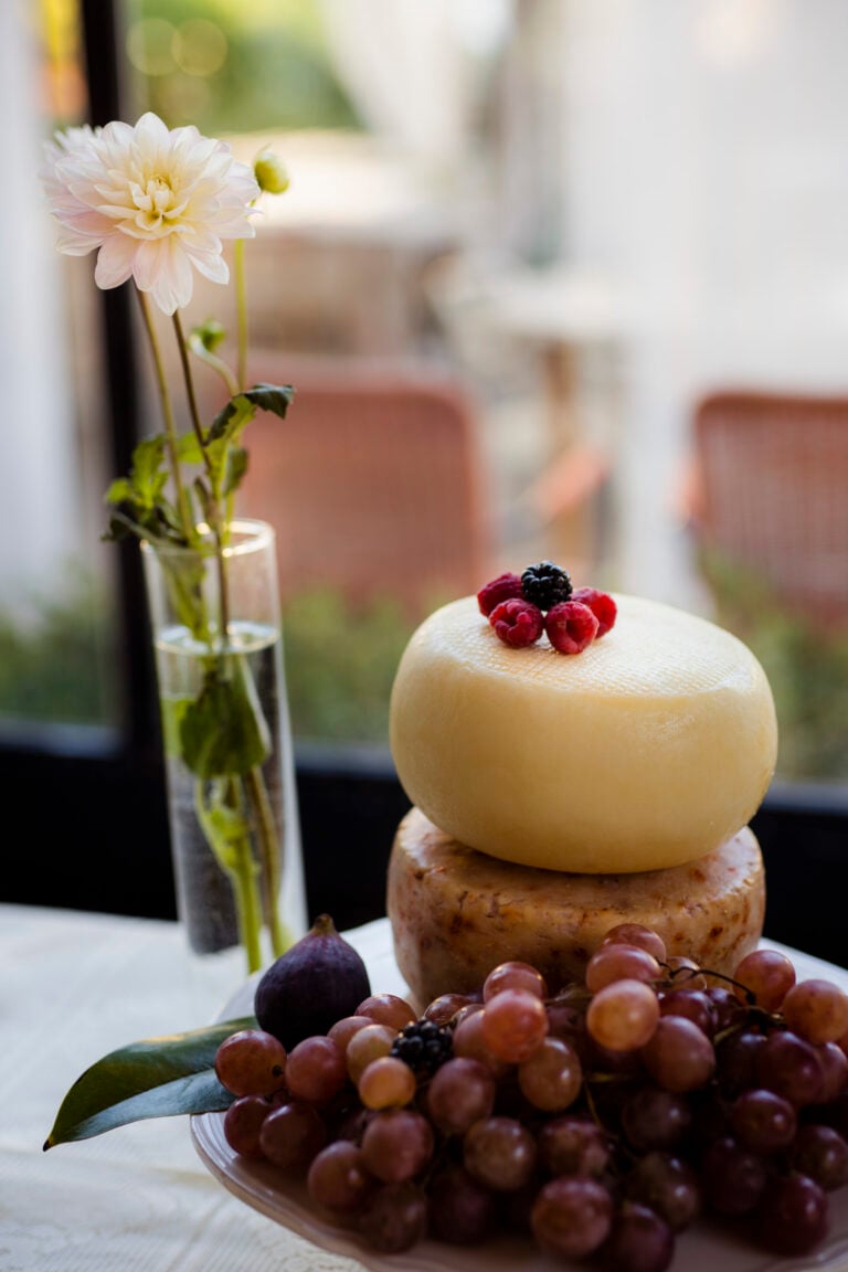 frutti fi bosco su due forme di formaggio con uva e un fico sul fondo e un fiore dentro ad un vaso di acqua, su tavolo con un centrino bianco
