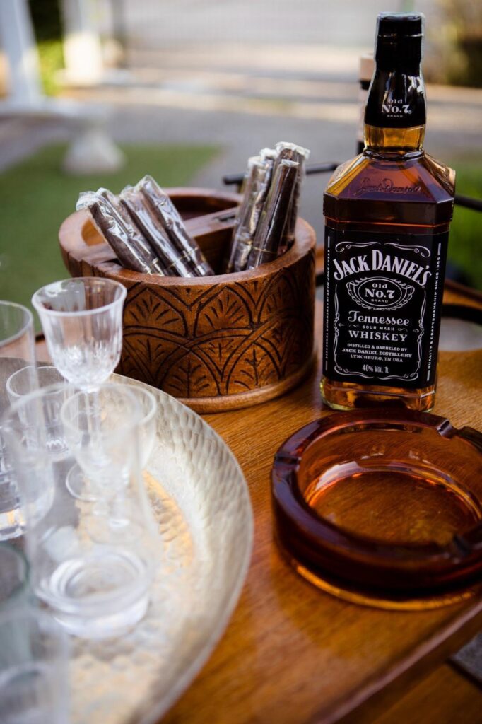 bottiglia di jack daniels con un posacenere scuro di fronte, sul lato sinistro della bottiglia dei sigari in un contenitore di legno rotondo intarsiato e sulla sinistra in basso un vassoio con dei calici vintage il tutto su un tavolino di legno